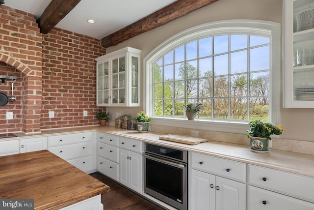 kitchen featuring oven, beam ceiling, brick wall, white cabinets, and glass insert cabinets