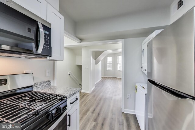 kitchen featuring appliances with stainless steel finishes, stacked washing machine and dryer, light wood-type flooring, white cabinetry, and baseboards