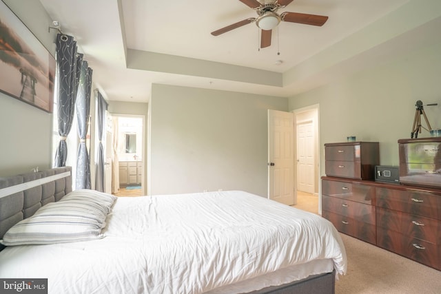 bedroom with light colored carpet, connected bathroom, a tray ceiling, and a ceiling fan