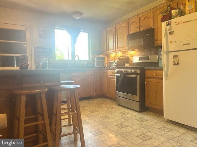 kitchen featuring freestanding refrigerator, stainless steel range with gas cooktop, black microwave, backsplash, and brown cabinetry