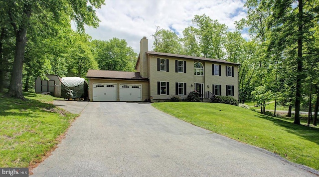 colonial-style house featuring a garage, asphalt driveway, a chimney, a storage shed, and a front lawn