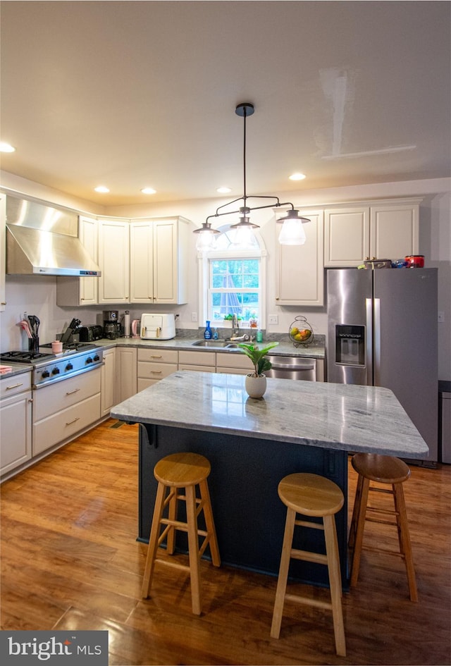 kitchen with wall chimney range hood, stainless steel appliances, a sink, a kitchen breakfast bar, and white cabinetry