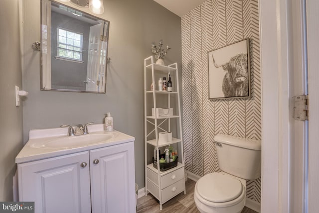 bathroom featuring an accent wall, wallpapered walls, toilet, vanity, and wood finished floors