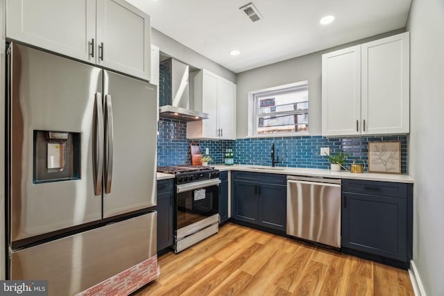 kitchen with appliances with stainless steel finishes, wall chimney range hood, light countertops, light wood finished floors, and a sink