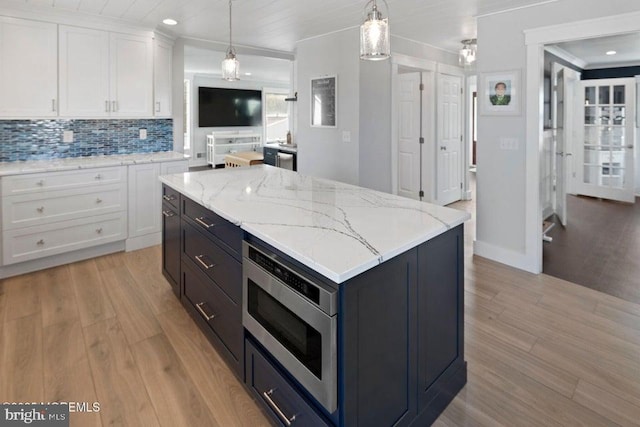 kitchen with white cabinets, stainless steel microwave, light wood-style floors, tasteful backsplash, and decorative light fixtures