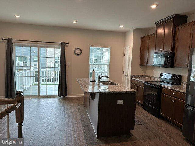 kitchen with black appliances, a sink, dark wood-type flooring, stone counters, and a center island with sink