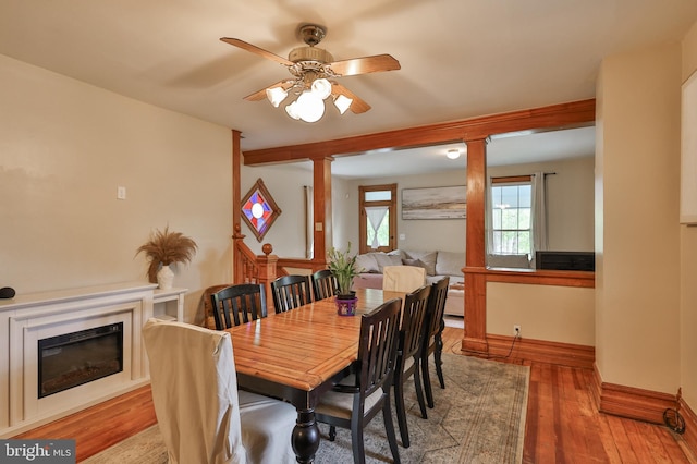 dining space with a ceiling fan, a glass covered fireplace, wood finished floors, baseboards, and decorative columns