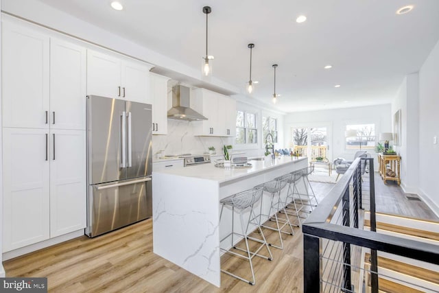 kitchen with stainless steel appliances, wall chimney range hood, light wood-style flooring, white cabinets, and an island with sink