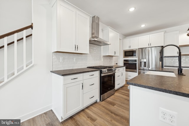 kitchen with appliances with stainless steel finishes, wall chimney exhaust hood, a sink, dark countertops, and recessed lighting
