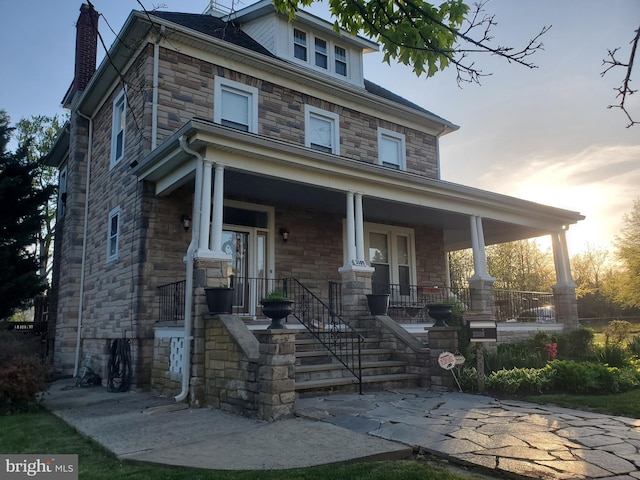 american foursquare style home with a chimney, stone siding, and covered porch