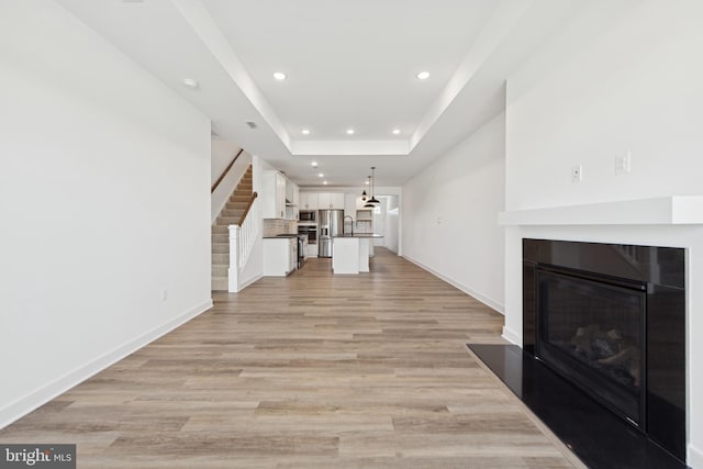 unfurnished living room with recessed lighting, a glass covered fireplace, stairs, a tray ceiling, and light wood-style flooring