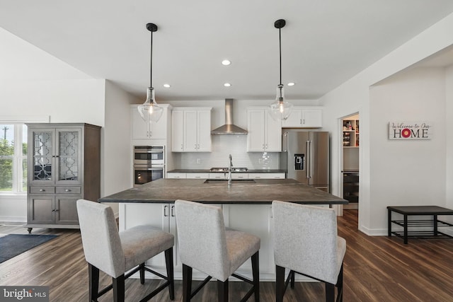 kitchen featuring wall chimney exhaust hood, stainless steel appliances, dark countertops, recessed lighting, and an island with sink