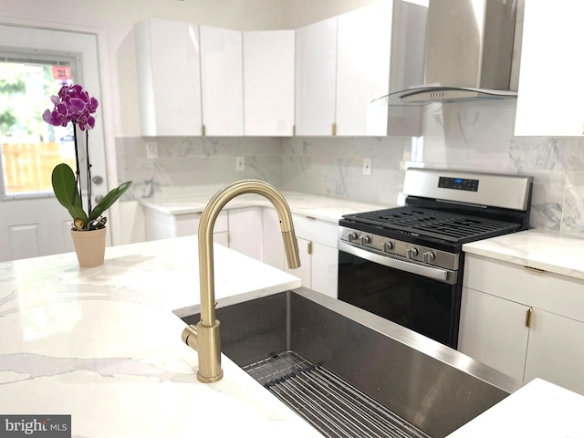 kitchen featuring wall chimney exhaust hood, gas range, white cabinets, decorative backsplash, and light stone countertops