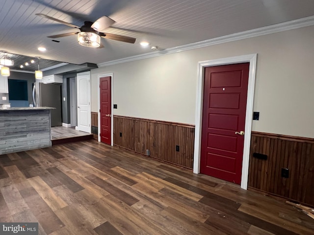 unfurnished living room with wainscoting, a ceiling fan, ornamental molding, dark wood-style flooring, and wooden walls