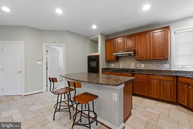 kitchen featuring under cabinet range hood, black appliances, a kitchen breakfast bar, a center island, and dark stone counters