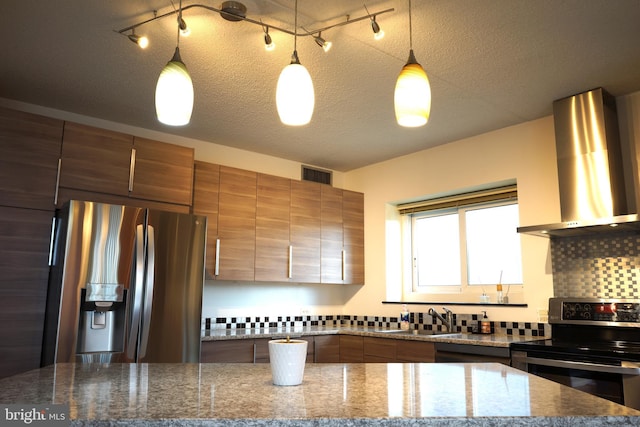 kitchen with stainless steel appliances, wall chimney range hood, a sink, brown cabinetry, and backsplash