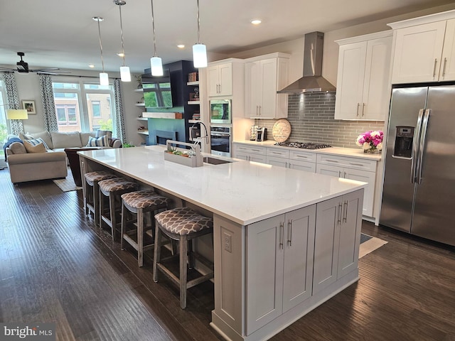 kitchen with appliances with stainless steel finishes, wall chimney exhaust hood, dark wood finished floors, open floor plan, and a center island with sink