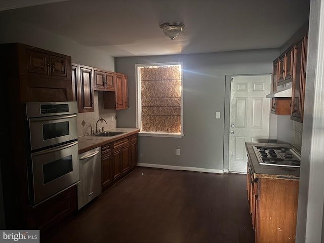 kitchen with stainless steel appliances, a sink, under cabinet range hood, baseboards, and tasteful backsplash