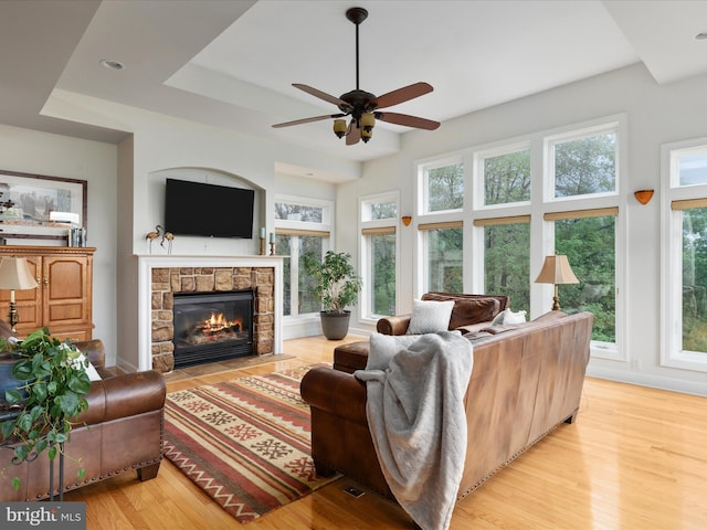 living area with ceiling fan, light wood finished floors, a stone fireplace, a raised ceiling, and recessed lighting