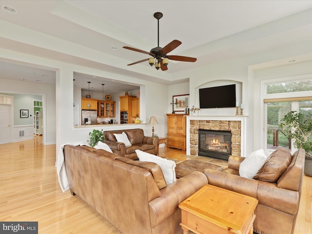 living room featuring light wood-type flooring, a ceiling fan, a fireplace, a raised ceiling, and a wainscoted wall