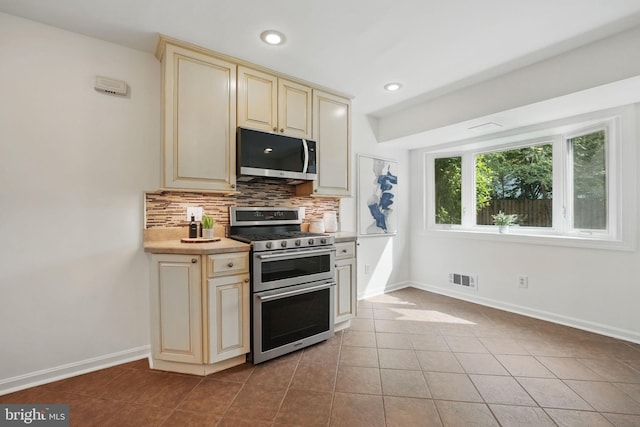kitchen featuring stainless steel appliances, cream cabinetry, tasteful backsplash, baseboards, and light countertops