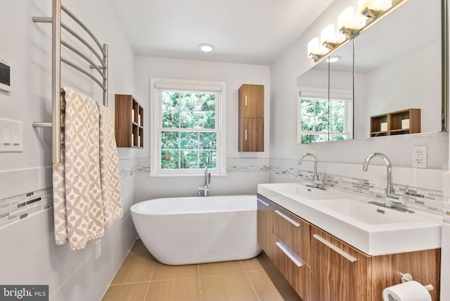 full bath with a soaking tub, tile patterned floors, tile walls, double vanity, and a towel warmer