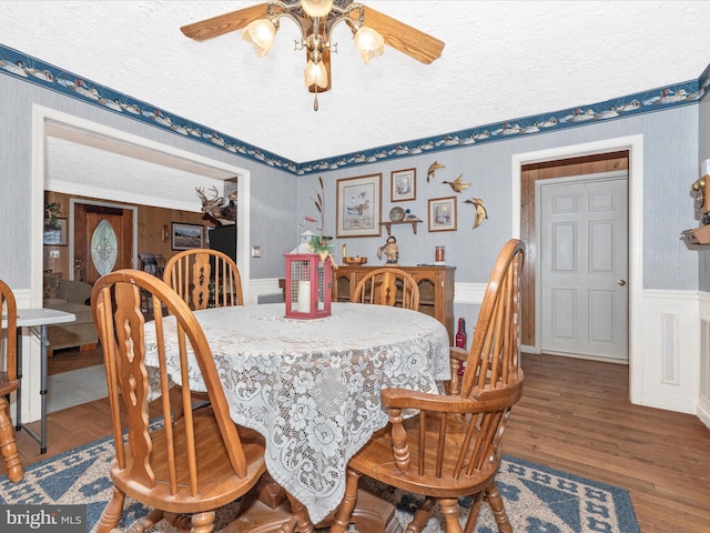 dining space with ceiling fan, wood finished floors, a wainscoted wall, a textured ceiling, and wallpapered walls