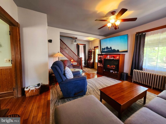 living room with radiator, a ceiling fan, stairs, wood finished floors, and a glass covered fireplace