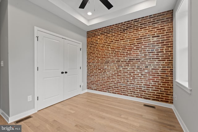 unfurnished bedroom with brick wall, a closet, baseboards, light wood-type flooring, and a tray ceiling