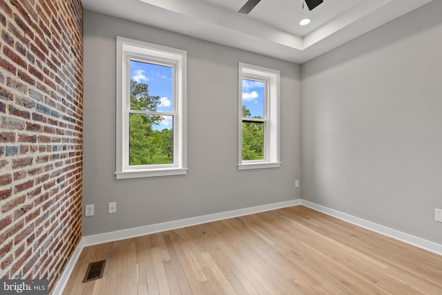 spare room featuring light wood finished floors, a ceiling fan, baseboards, brick wall, and a raised ceiling