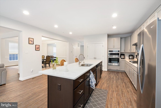 kitchen featuring appliances with stainless steel finishes, a sink, light countertops, a center island with sink, and dark wood-style floors