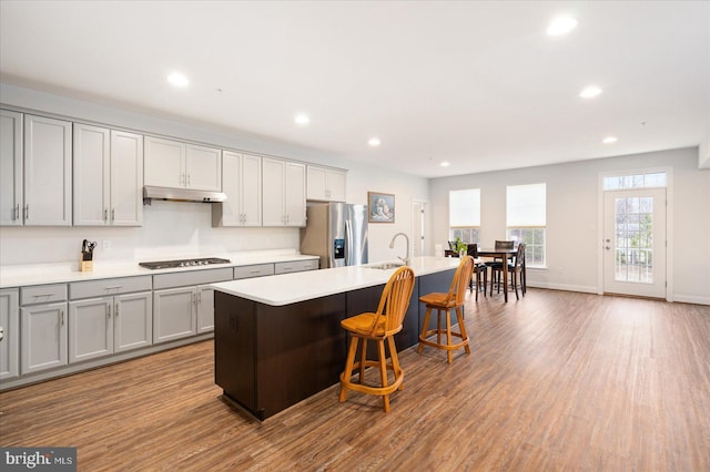 kitchen featuring stainless steel fridge, gray cabinets, under cabinet range hood, a sink, and an island with sink