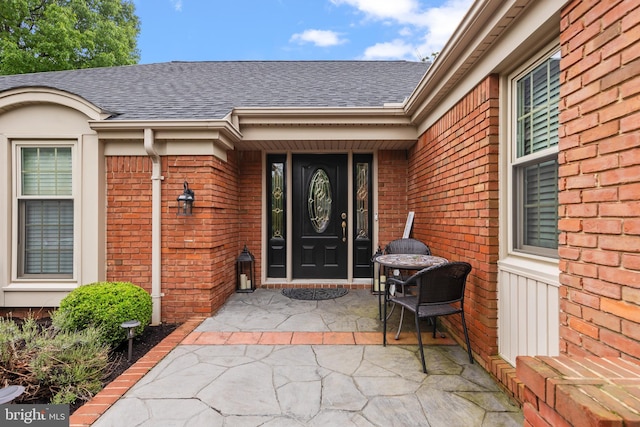 property entrance with roof with shingles, brick siding, and a patio area