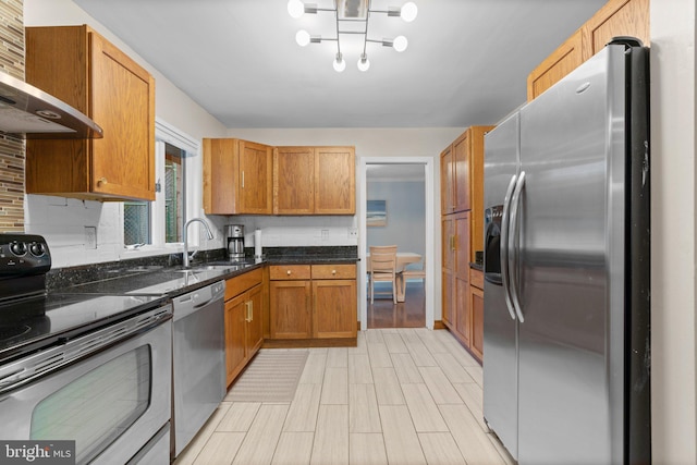 kitchen with stainless steel appliances, wall chimney exhaust hood, a sink, dark stone counters, and brown cabinetry