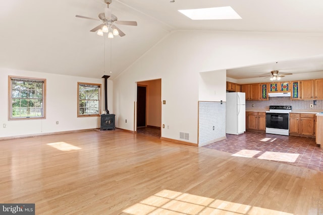 unfurnished living room with ceiling fan, a wood stove, a skylight, light wood-style floors, and high vaulted ceiling