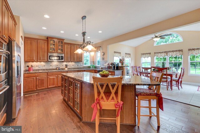 kitchen with stainless steel appliances, brown cabinetry, a kitchen island, glass insert cabinets, and a kitchen breakfast bar