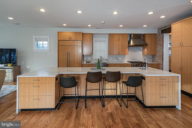 kitchen with built in refrigerator, wall chimney range hood, a large island with sink, wood finished floors, and modern cabinets