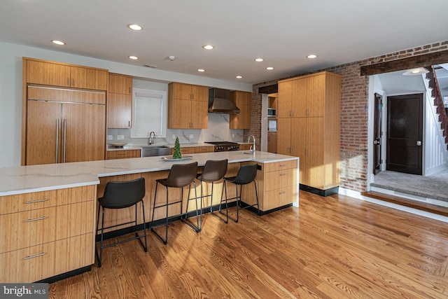 kitchen with built in refrigerator, wall chimney range hood, wood finished floors, a breakfast bar area, and recessed lighting