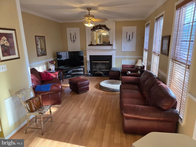 living area with wood finished floors, a ceiling fan, ornamental molding, a fireplace with flush hearth, and baseboards