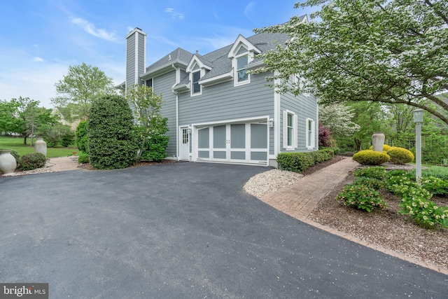 view of side of property featuring asphalt driveway, a chimney, a garage, and a shingled roof