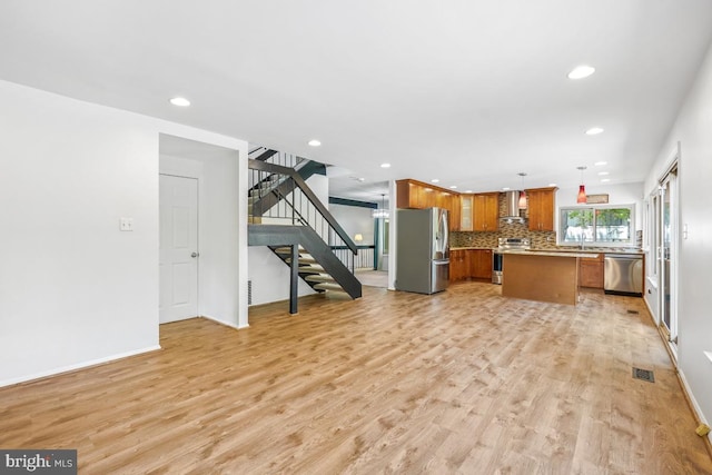 kitchen featuring stainless steel appliances, wall chimney range hood, brown cabinetry, open floor plan, and light wood-style floors