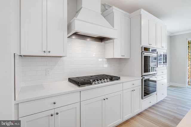 kitchen featuring custom exhaust hood, appliances with stainless steel finishes, white cabinets, decorative backsplash, and light wood finished floors