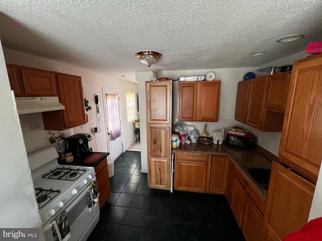 kitchen with white range with gas cooktop, under cabinet range hood, a textured ceiling, dark tile patterned floors, and brown cabinets