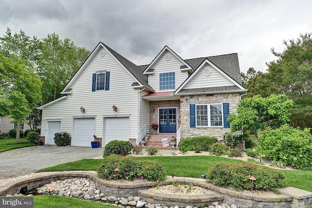 traditional-style house featuring a garage, decorative driveway, stone siding, a front yard, and a shingled roof