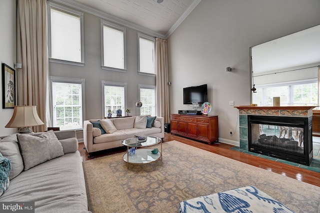 living area with ornamental molding, a towering ceiling, wood finished floors, a tiled fireplace, and plenty of natural light