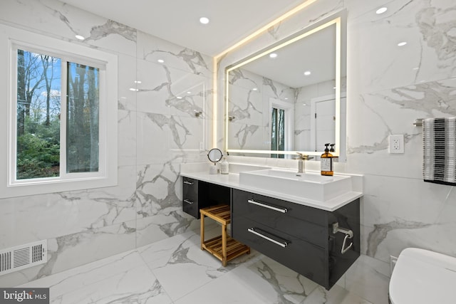 bathroom with toilet, vanity, tile walls, marble tiled flooring, and stone wall