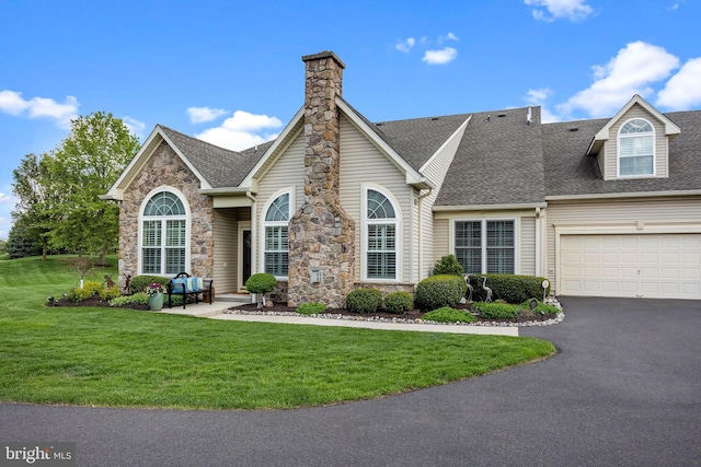 traditional-style house featuring driveway, a front yard, roof with shingles, and stone siding