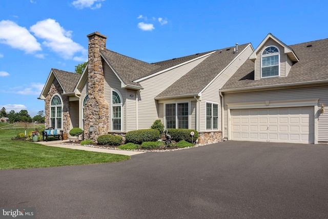 view of front facade featuring stone siding, roof with shingles, asphalt driveway, a front yard, and a garage
