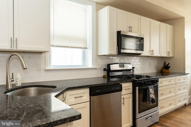 kitchen featuring appliances with stainless steel finishes, a sink, tasteful backsplash, dark stone counters, and white cabinets