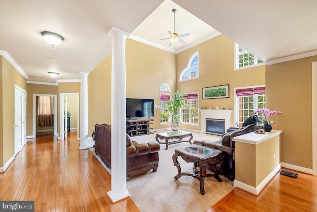 living room featuring decorative columns, light wood-style flooring, crown molding, a glass covered fireplace, and baseboards